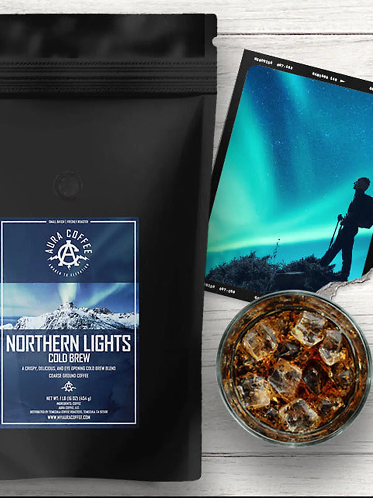 A black bag of "Northern Lights Cold Brew" coffee with a label featuring a mountain landscape under the Northern Lights, next to a glass of iced coffee and a postcard with a similar aurora borealis theme.