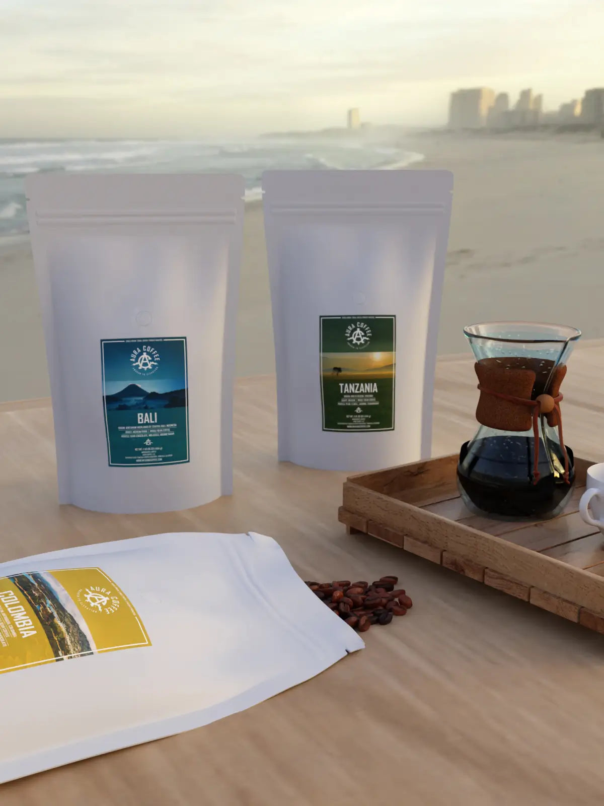 White pouches of Aura Coffee Studio's single-origin coffees, including Bali and Tanzania, displayed on a wooden surface with a Chemex coffee maker, a cup of coffee, and coffee beans, set against a blurred beach and ocean background.